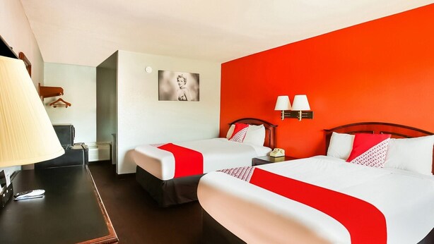 A hotel room with two beds, a desk with a lamp, a coat rack, and a wall-mounted picture.