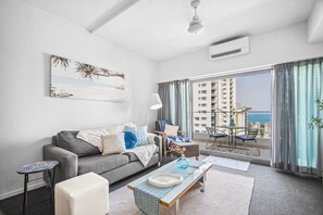 Un salon moderne avec un canapé gris, une petite table, un balcon offrant une vue sur les immeubles de grande hauteur et un ventilateur de plafond.