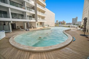 Une piscine sur le toit avec une terrasse en bois, un bâtiment blanc doté de balcons et un ciel d’un bleu limpide.