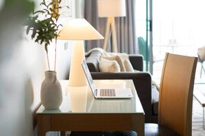 Ein Laptop steht auf einem Glastisch, dazu eine Vase mit einer Pflanze, eine Lampe, ein Sofa und ein Sessel in einem gut beleuchteten Raum.