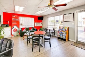 Une chambre d’hôtel avec un mur d’accent rouge, un ventilateur de plafond, un coin repas avec une table ronde et des chaises, un canapé, une télévision et une fenêtre avec des stores.