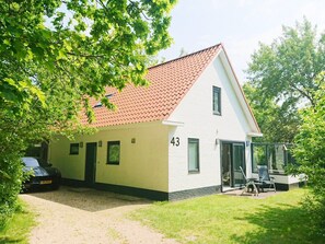 A two-story house with a red-tiled roof, a number '43' on the side, a car parked in the driveway, and a glass door leading to a covered outdoor area. - Park Retreat With Fireplace (Koudekerke)