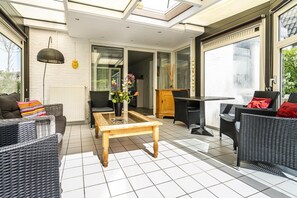 Eine sonnendurchflutete Terrasse mit Korbmöbeln, einem Tisch mit Glasplatte und Blick durch eine Glastür.