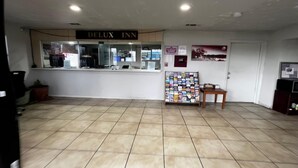 Uma área de recepção de hotel com uma placa indicando 'DELUXE INN', um balcão de atendimento, um mural de avisos e um piso de azulejos.