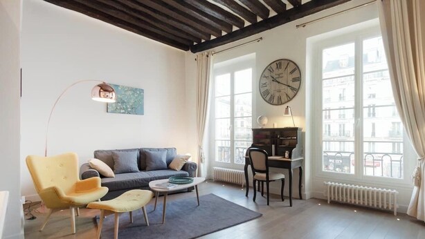 A living room with a sofa, armchairs, a coffee table, a clock, and a view of buildings through large windows. - Charming high ceiling Le Marais St Paul apartment (Paris)