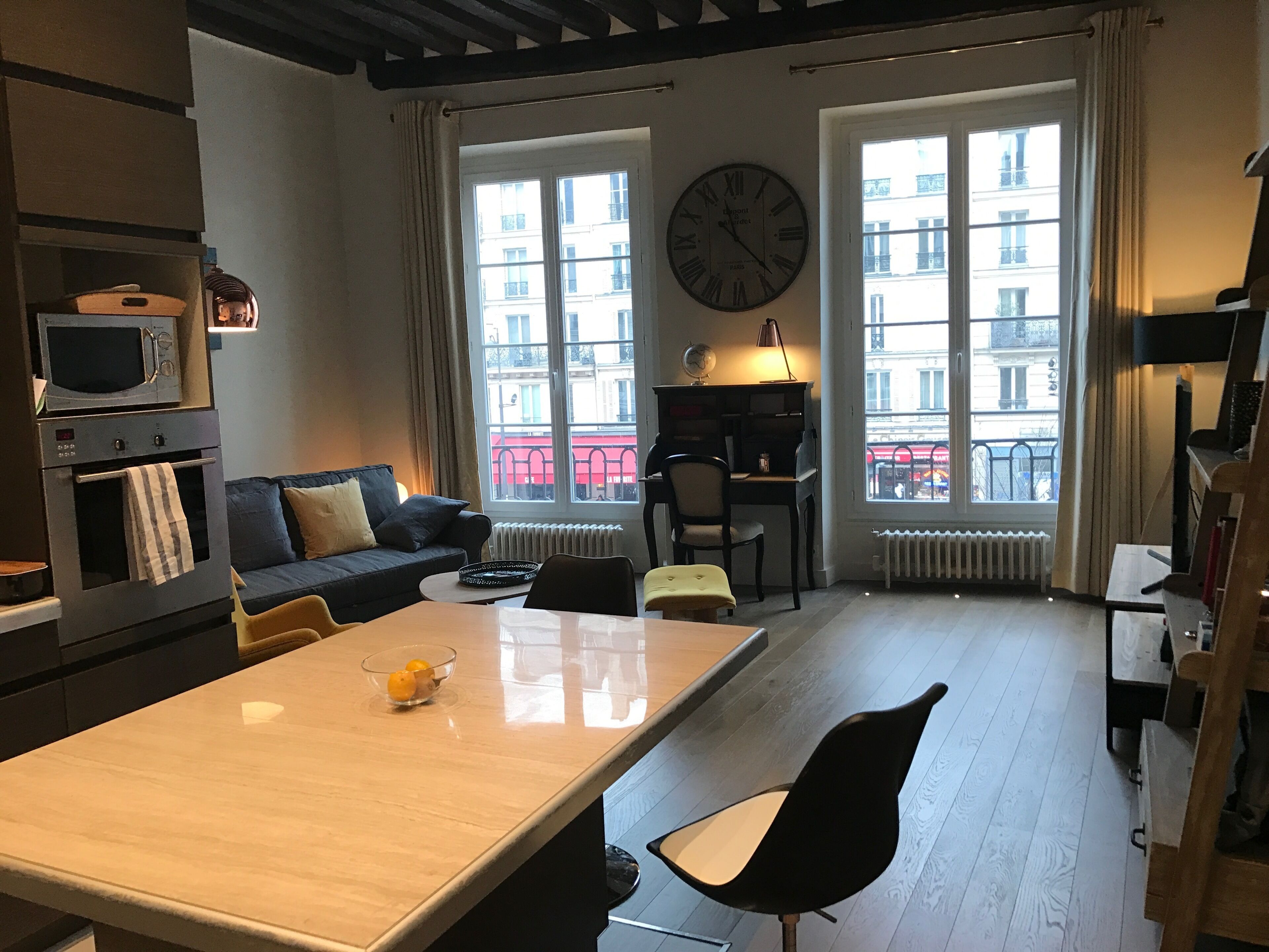 A modern living room with a sofa, a dining table, a black chair, a clock, and a view of buildings through large windows.