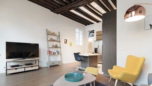 A modern living room with a flat-screen TV, a coffee table, a yellow armchair, and a bookshelf. - Charming high ceiling Le Marais St Paul apartment (Paris)