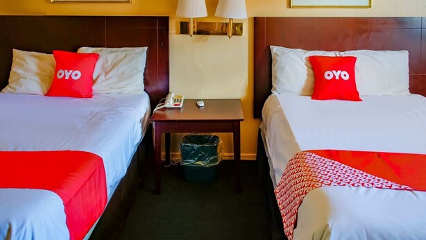 A hotel room with two beds, each with a red pillow labeled 'OYO,' a bedside table with a phone and lamp, and a trash bin.