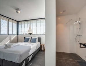 Ein modernes Hotelzimmer mit Bett, kleinem Tisch, einer Segeltuch-Dekoration, einer Dusche und großen Fenstern.