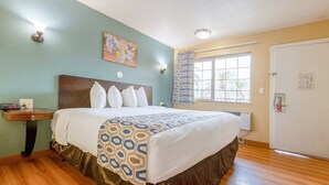 A hotel room with a bed, bedside table, wall art, window with curtains, and a door. - King Bed Non-Smoking (Oceanside, CA)