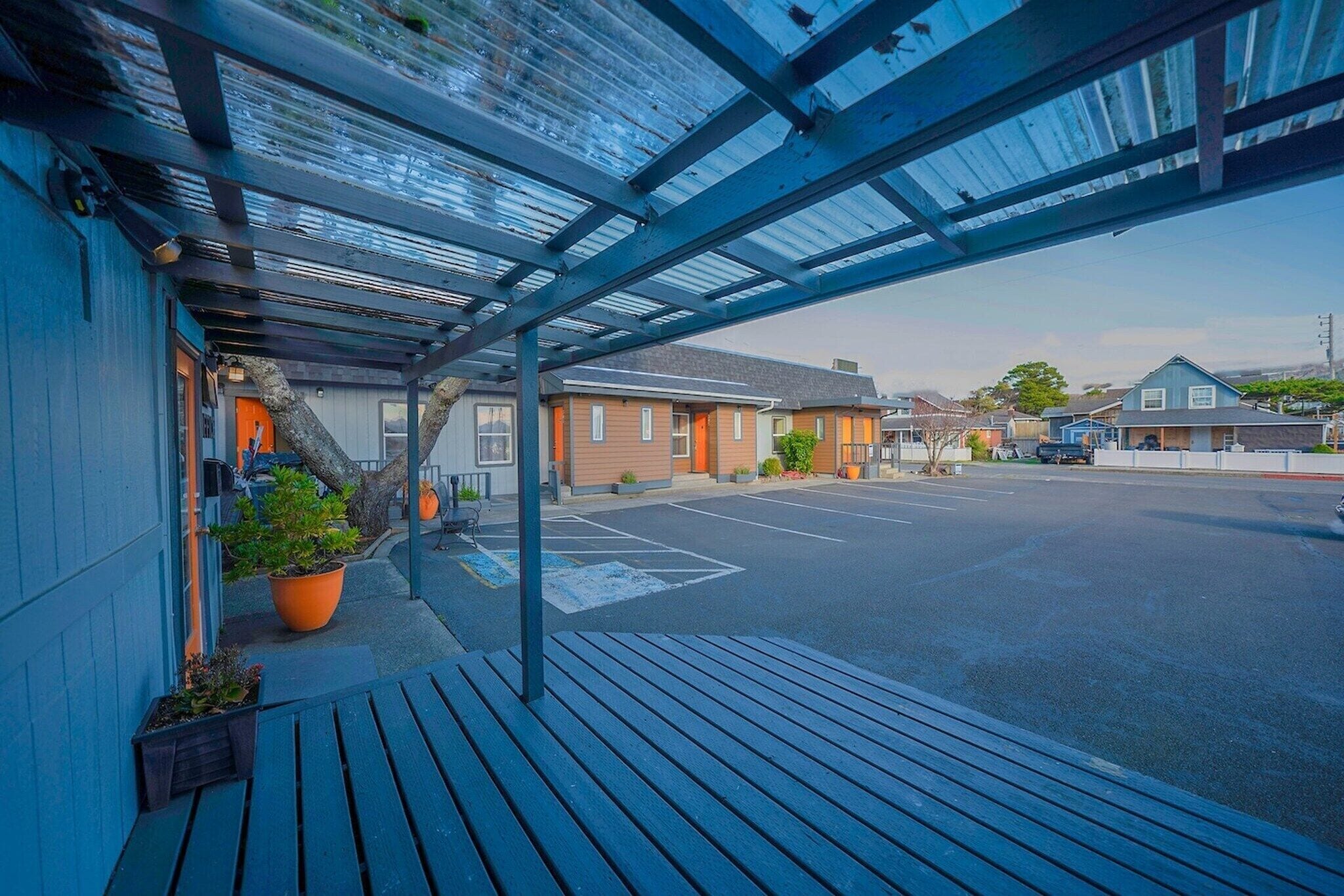 A covered outdoor area with a wooden bench, potted plants, and a parking lot.