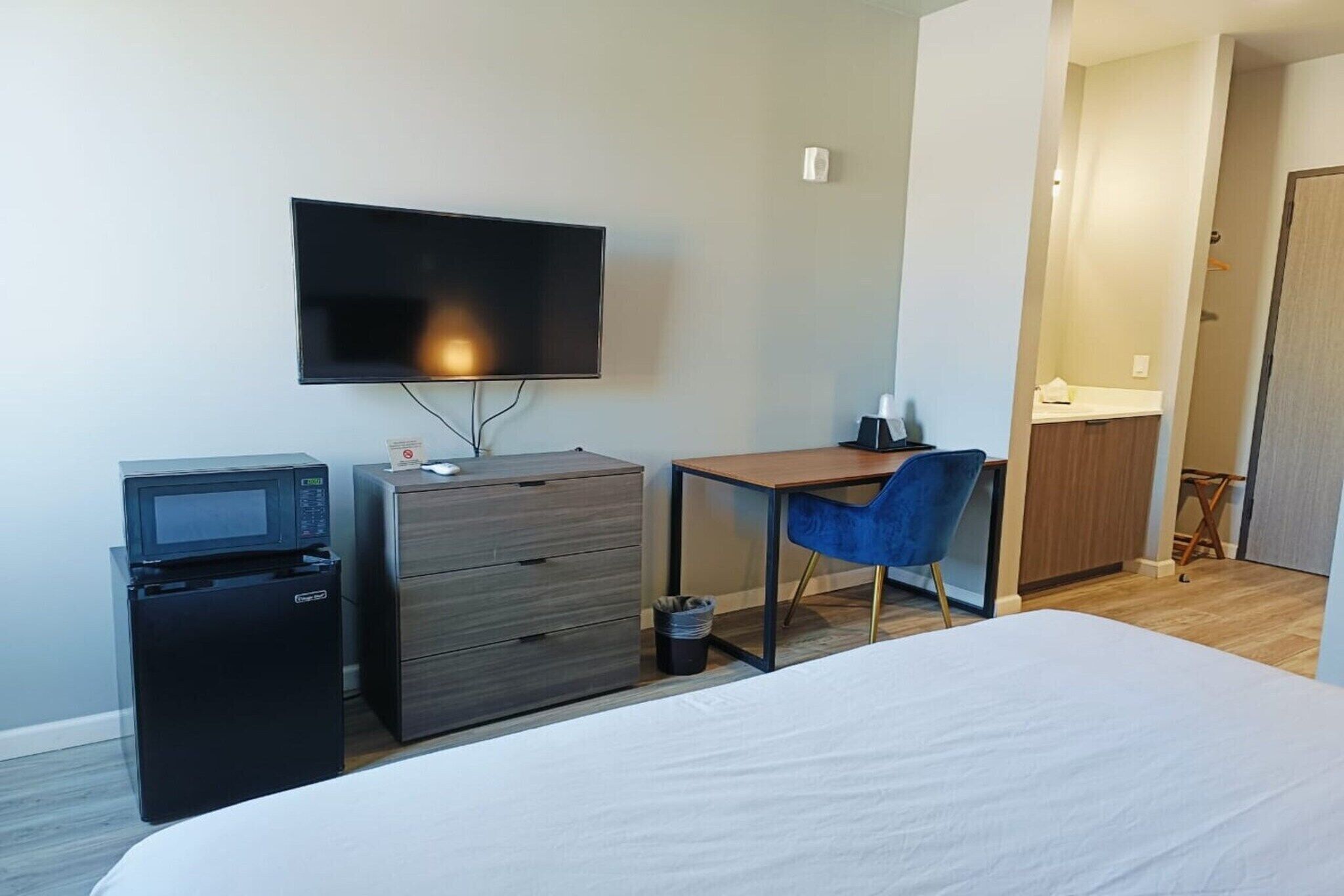 Habitación de hotel con una cama, un escritorio con silla, televisor, microondas y un pequeño cubo de basura.