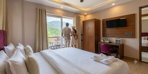 Quarto de hotel com uma cama grande, televisão fixada na parede, armário de madeira e varanda com vista para as montanhas.