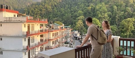 A balcony with a view of a mountainous landscape, a building with multiple floors, and a table with a cup.