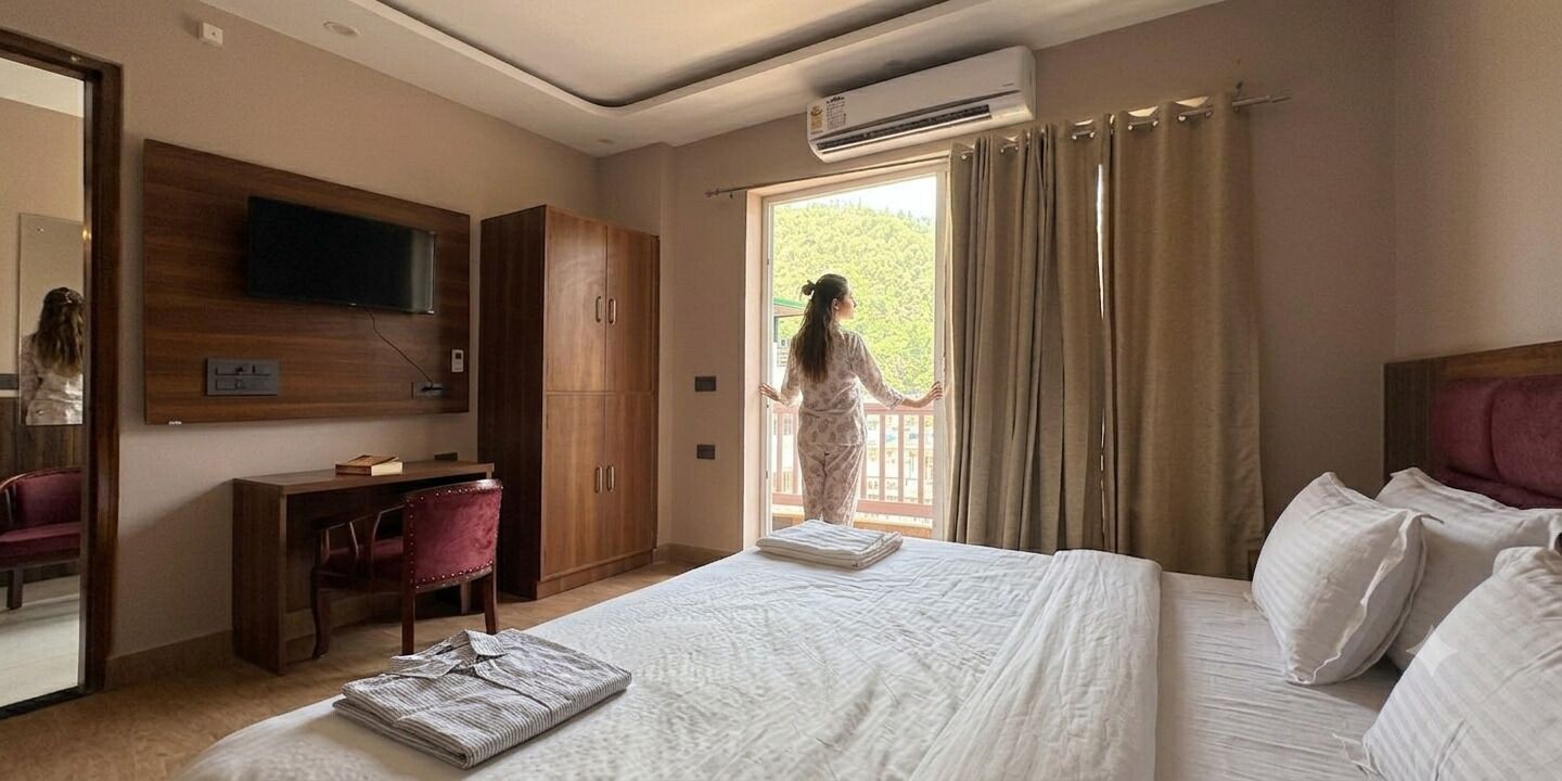 A hotel room with a bed, a desk, a chair, a wardrobe, a television, and an air conditioning unit.