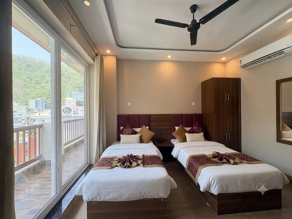 A hotel room with two beds, a ceiling fan, and a balcony with a view of buildings and greenery.