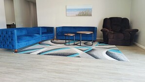 Ein Zimmer mit einem blauen Ecksofa, einem kleinen runden Tisch, einem Liegesessel und einem gemusterten Teppich.