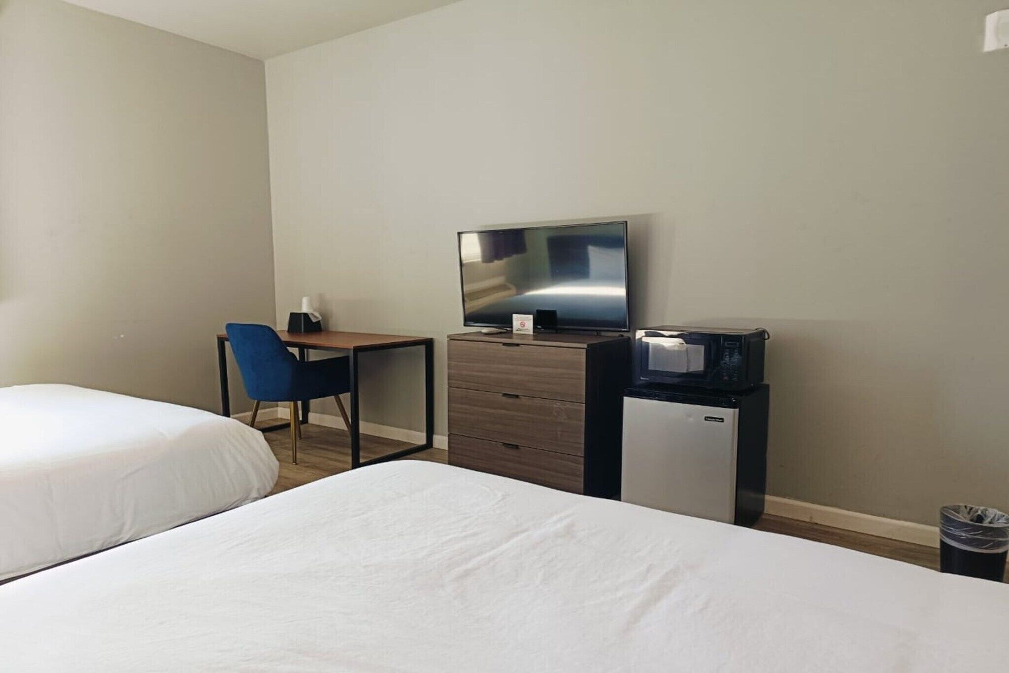Habitación de hotel con dos camas, un escritorio con silla, televisor, mini refrigerador y un cesto de basura.