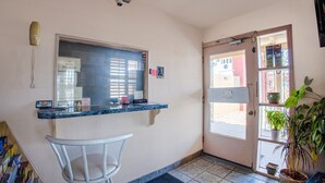 Un mostrador de recepción con un teléfono, una computadora y una silla. Hay una puerta con un letrero y una ventana con persianas.