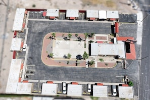 Vista aérea de un estacionamiento con múltiples espacios, un área central ajardinada con árboles y edificios adyacentes con techos rojos.