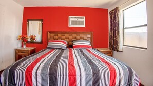 Una habitación con pared roja, cabecera de madera, cama grande con ropa de cama a rayas, un espejo, una ventana con cortinas y una pequeña mesa con un jarrón de flores.