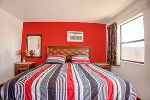 Una habitación con pared roja, cabecera de madera, cama grande con ropa de cama a rayas, un espejo, una ventana con cortinas y una pequeña mesa con un jarrón de flores.