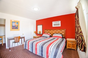 Habitación de hotel con cama, escritorio, silla, mesita de noche y aire acondicionado de pared.