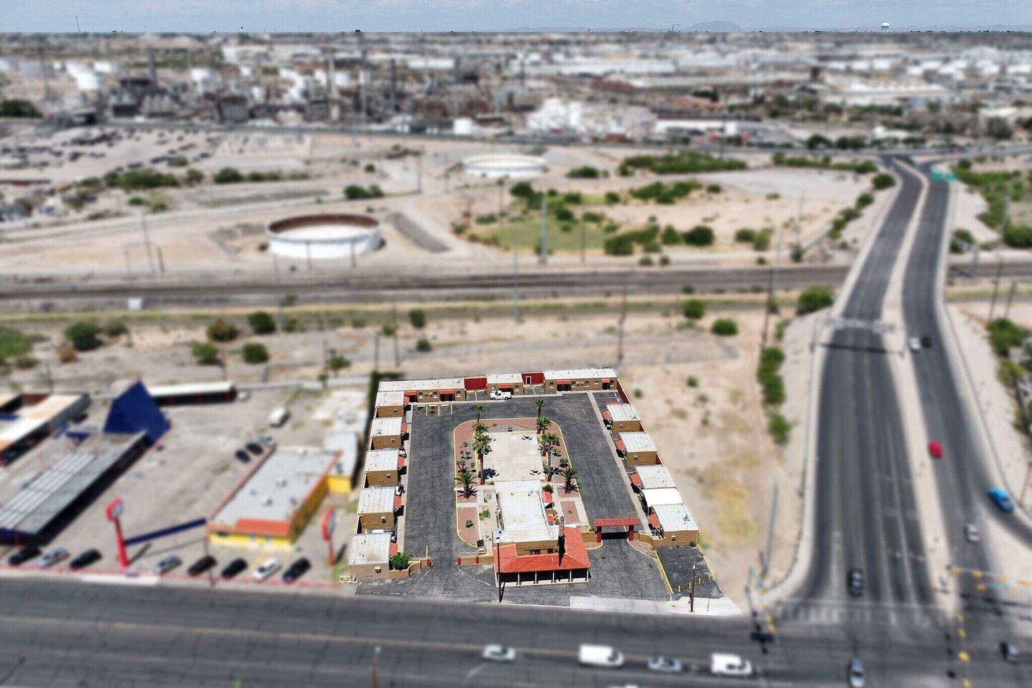 Une vue aérienne d’un parking avec plusieurs rangées de voitures garées, une zone centrale dégagée et une route qui s’éloigne du parking.