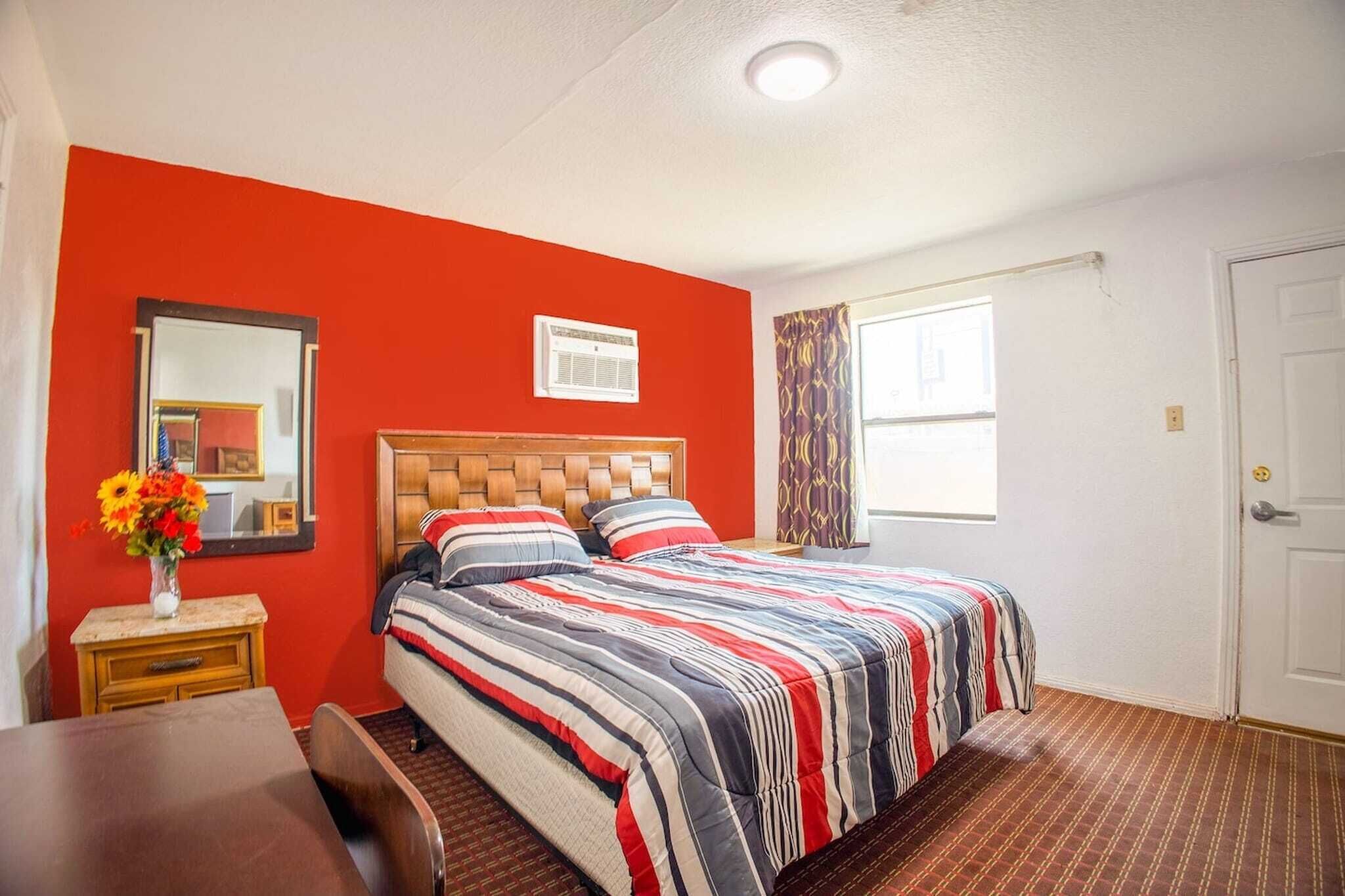 A hotel room with a bed, a desk, a chair, a window with curtains, a mirror, and a door.