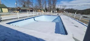 Une piscine extérieure recouverte de neige, dotée d’une terrasse en bois et d’une clôture blanche.