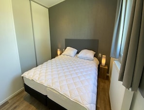 A neatly made bed with a quilted white duvet, two bedside lamps, and a grey headboard in a room with wooden flooring and a grey wall. - Beachside Chalet Near De Pluimpot for 4 (Scherpenisse)