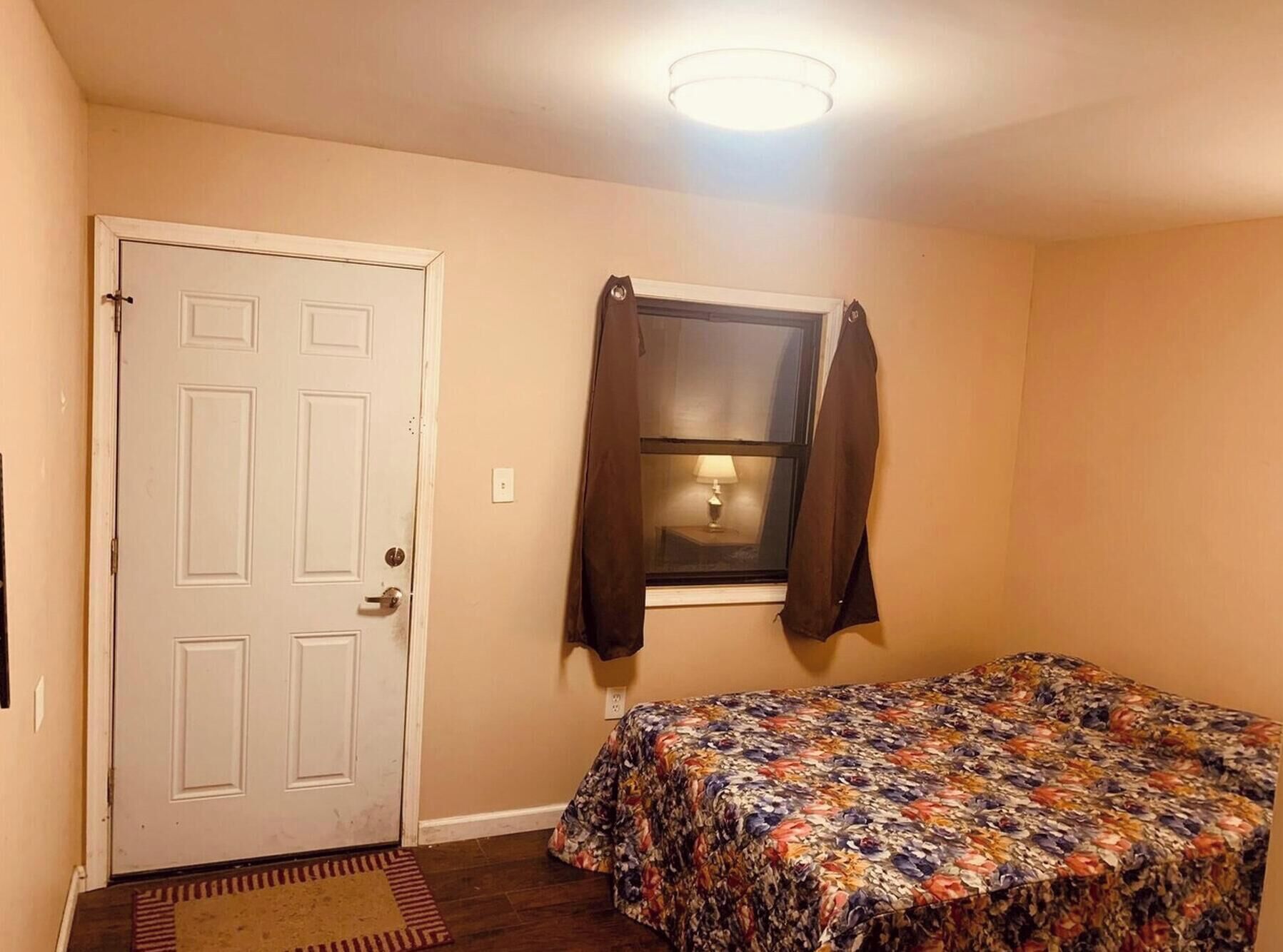 A bedroom with a door, a window with curtains, a bed with a floral bedspread, and a ceiling light.