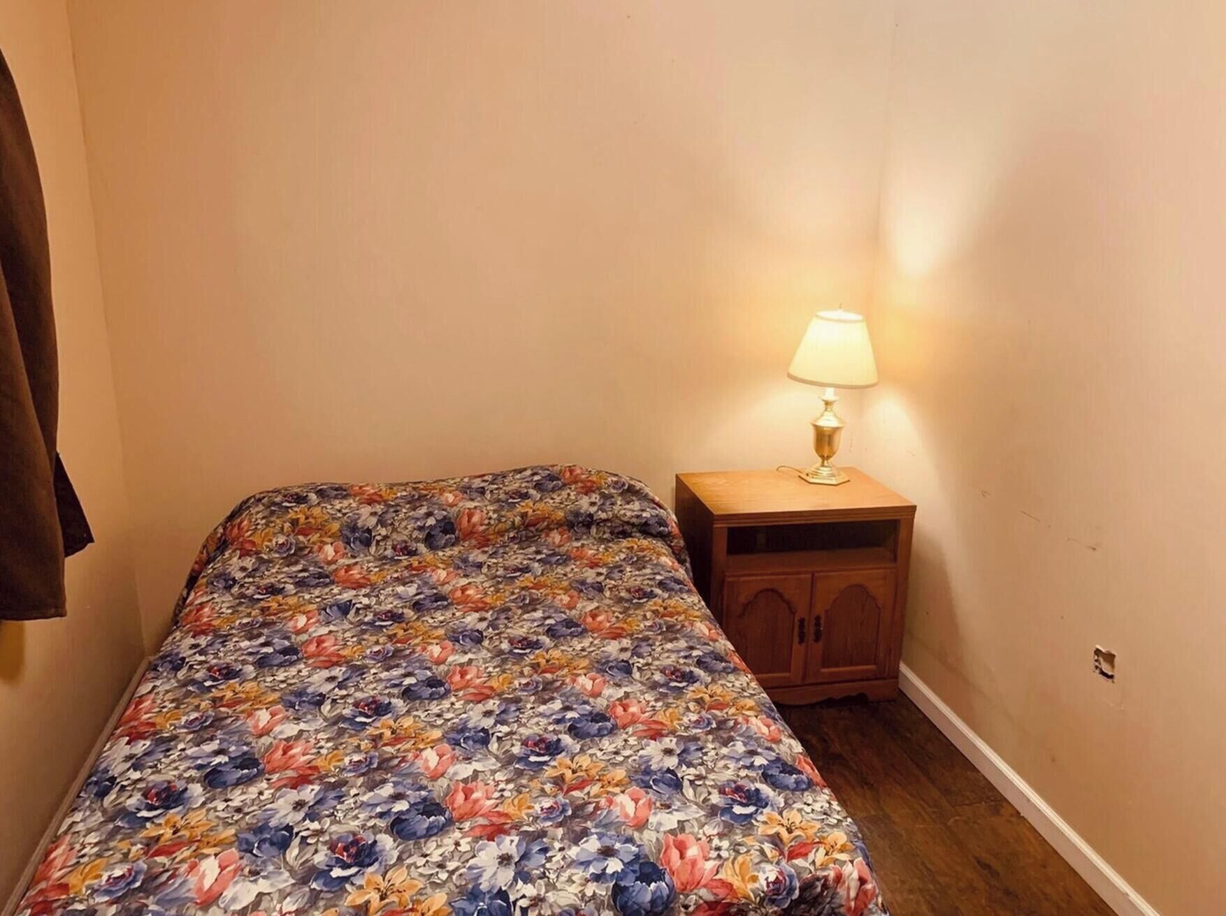 Une petite chambre avec une couvre-lit à motifs floraux, une table de chevet en bois avec une lampe, et un manteau marron accroché au mur.