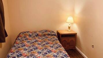 Une petite chambre avec une couvre-lit à motifs floraux, une table de chevet en bois avec une lampe, et un manteau marron accroché au mur.