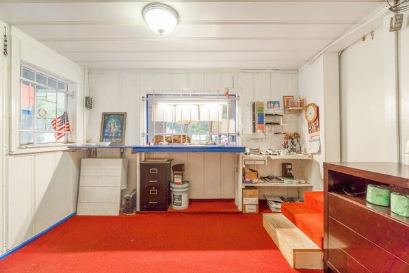 A compact room with a red carpet, a window with an American flag, a desk with a lamp, and a clock on the wall.