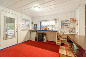 A room with a red carpet, white walls, a door with a mesh screen, a window with blinds, a desk with a lamp, and shelves with various items.
