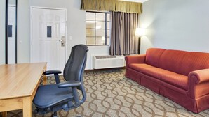 Habitación de hotel con un escritorio de madera, una silla de oficina negra, un sofá rojo, una ventana con cortinas y un aire acondicionado instalado en la pared.