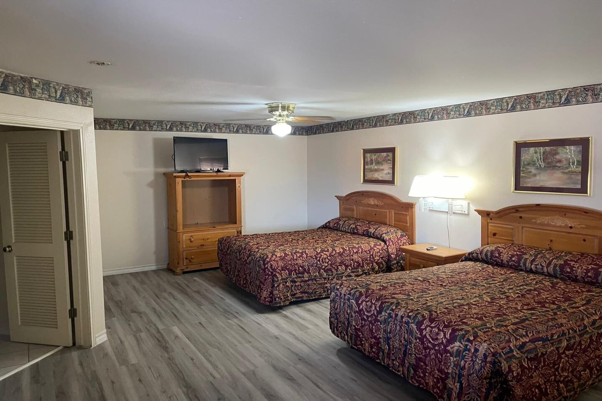 A bedroom with two beds, a TV on a wooden stand, a ceiling fan, and a lamp.