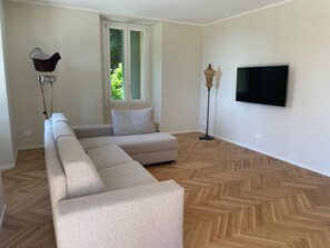 A living room with a sectional sofa, a floor lamp, a television mounted on the wall, and a window with a view of greenery.
