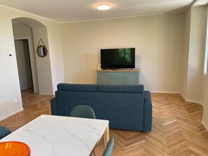 A living room with a sofa, a television on a cabinet, a dining table with chairs, and a herringbone-patterned floor. - Apartment 'The Mill' in Historic Villa near Lake Como (Caslino D'erba)