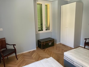 A room with a wooden herringbone floor, a window with blinds, a wardrobe, a small chest, and two chairs.