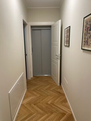 Käytävä, jossa on puinen kalanruotokuvioinen lattia, valkoiset seinät ja käytävän päässä oleva suljettu ovi.