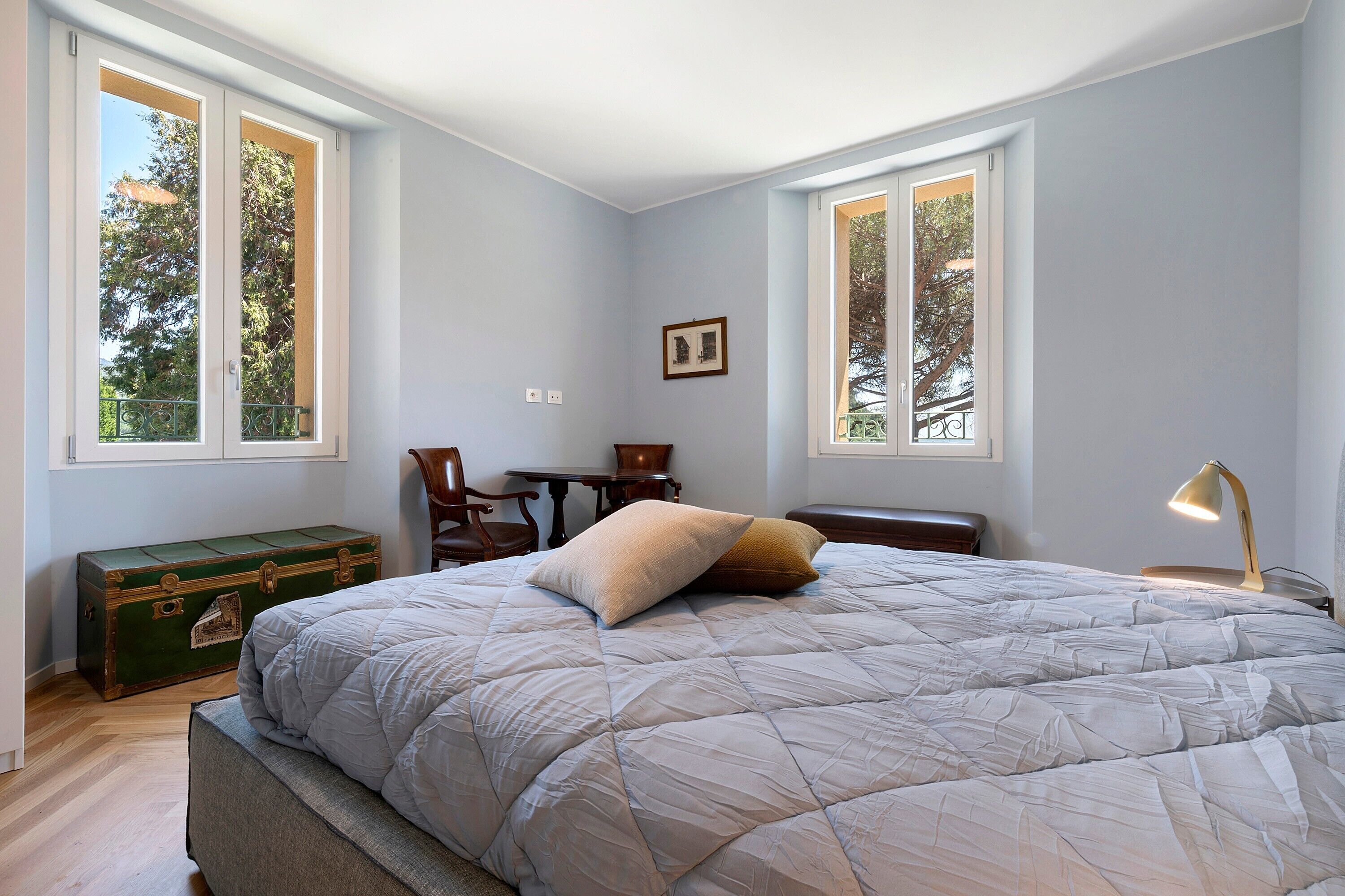 Ein Schlafzimmer mit einem großen Bett, einem Schreibtisch und einem Sessel. Zwei Fenster sorgen für natürliches Licht.