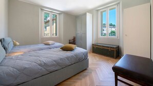Un dormitorio con cama, un banco, un aparador y dos ventanas con balcones.