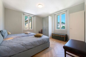 A bedroom with a bed, a bench, a chest of drawers, and two windows with balconies.