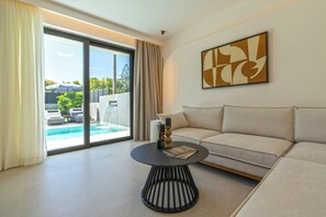 A modern living room with a sofa, a round coffee table, and a large window with a view of a pool and outdoor furniture. - Green villa II, Stylish villa with pool near beaches in Rethymno (Rethymno, Pigi)