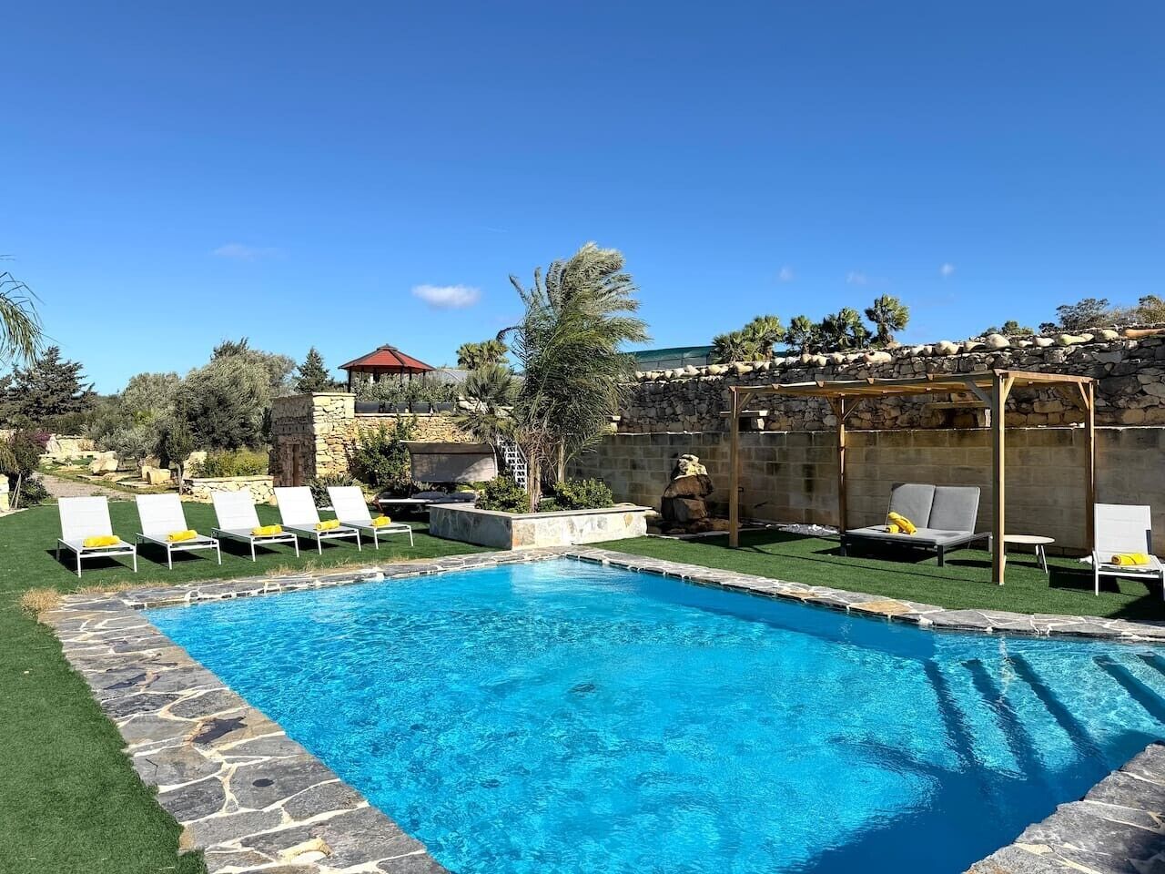A swimming pool with lounge chairs, a stone wall, and a pergola.