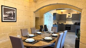 Een eetruimte met een ronde tafel voor zes personen, begrensd door stenen muren en een gewelfde deuropening naar een keuken.