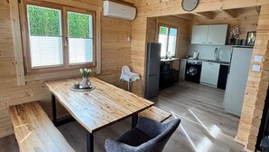 A wooden cabin interior with a dining table, bench, gray chair, kitchen area, and a clock on the wall.