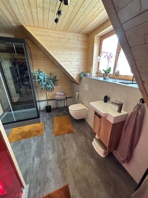 Ein modernes Badezimmer mit Holzboden, einer Glasdusche, einer Toilette, einem Waschbecken und einem Fenster mit Pflanzen.